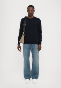Marineblå sweater, lyseblå denim jeans, hvide sneakers. Model bærer en beige trekantet taske med en sort skulderrem. Enkel, moderne design.