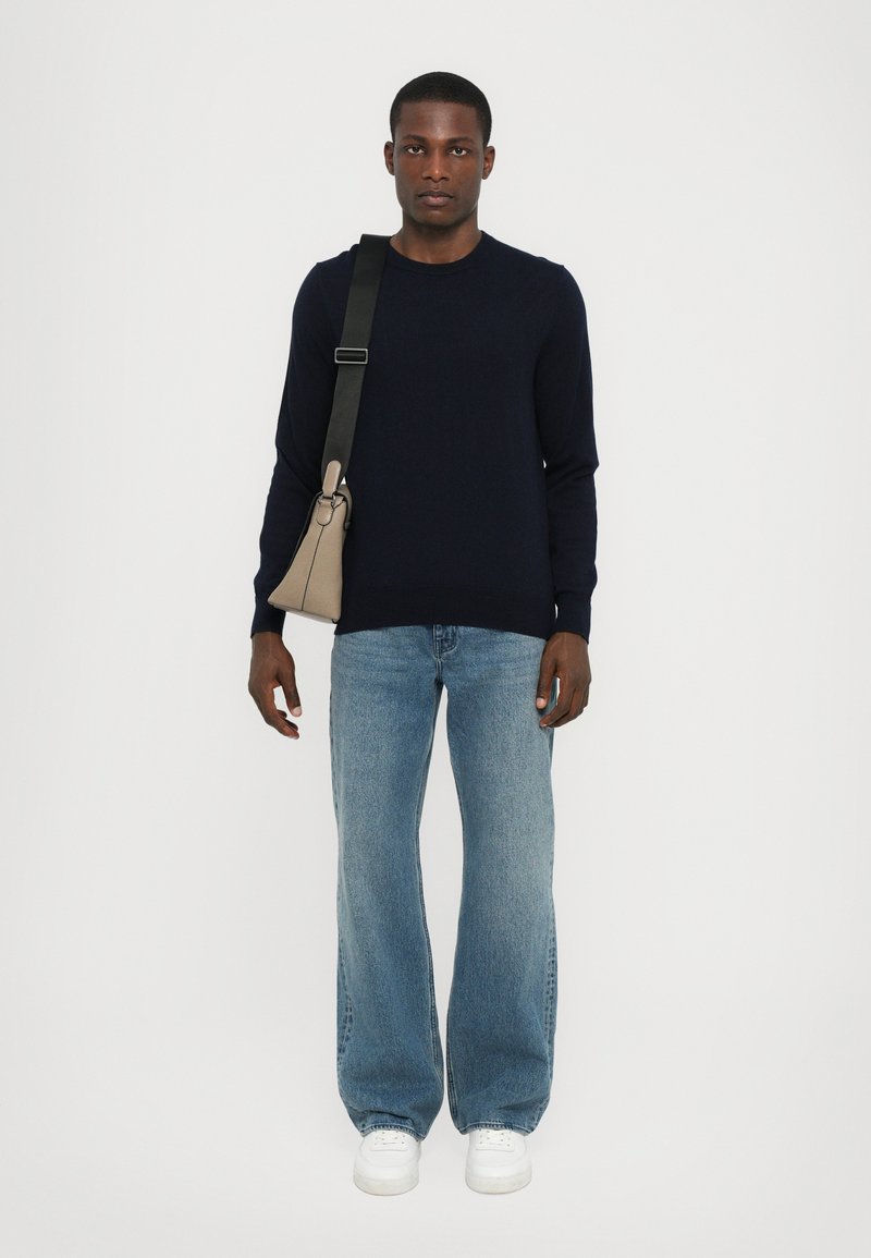 Marineblå sweater, lyseblå denim jeans, hvide sneakers. Model bærer en beige trekantet taske med en sort skulderrem. Enkel, moderne design.