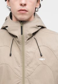Μπεζ μπουφάν με κουκούλα και φερμουάρ εμπρός, διακοσμημένο με κυματιστά μοτίβα και μαύρα κορδόνια. Το λογότυπο "OBEY" είναι κεντημένο με λευκό χρώμα.
