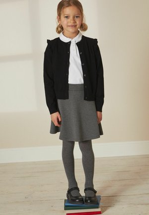 Ung pige står på stablede bøger iført en hvid polo, sort cardigan, grå nederdel, grå strømpebukser og sorte sko.