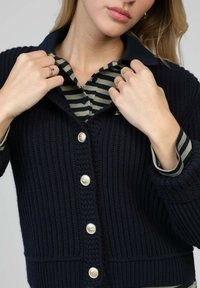 Cardigan en maille bleu marine avec un motif texturé, orné de grands boutons argentés. En dessous, une chemise à col rayée foncée est visible.
