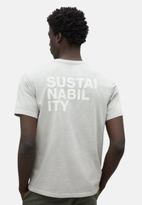 Camiseta gris de algodón, de manga corta, con un gran texto blanco "SOSTENIBILIDAD" en la parte de atrás. Diseño de cuello redondo.