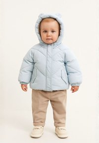 Világoskék párnázott dzseki medvefülekkel, cipzárral és két oldalsó zsebbel, bézs nadrággal és világos sportcipőkkel párosítva.