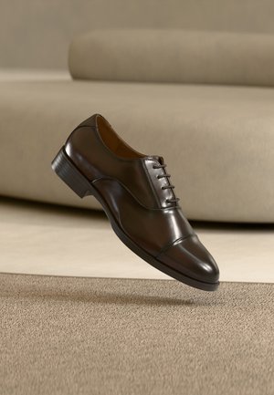 Błyszczący, ciemnobrązowy skórzany but wizytowy na sznurowadła, unoszący się nad beżowym dywanem przed beżową sofą w podobnym odcieniu.