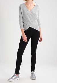 Femme portant un pull en maille gris en forme de cache-cœur, un pantalon skinny noir et des baskets argentées, debout devant un fond clair uni.