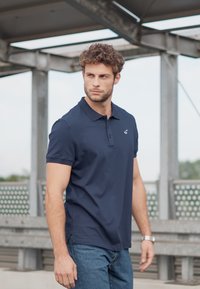 Marineblaues Poloshirt mit einer Knopfleiste mit zwei Knöpfen, kurzen Ärmeln und einem kleinen Logo. Kombiniert mit Jeans und einer silbernen Uhr. Lässige Umgebung.