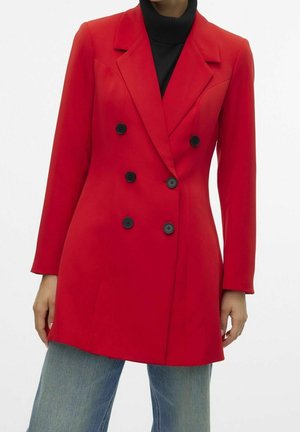 Femme portant un blazer rouge vif à double boutonnage avec des boutons noirs sur un col roulé noir et un jean bleu sur fond blanc.