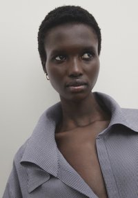 Chemise à rayures bleues et blanches avec un grand col, offrant une coupe décontractée. Le mannequin a les cheveux courts et porte une simple boucle d'oreille en or.