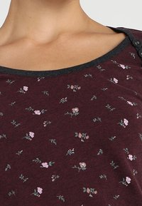 Bordeaux topp med ett mjukt, texturerat tyg. Har små rosa och gröna blommönster, en rundad halsringning och mörkgrå kant med knappar.