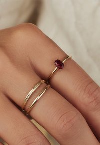 Des anneaux en or avec une finition polie, dont l'un présente une pierre précieuse rouge en forme d'ovale, reposant contre un fond de peau texturée.