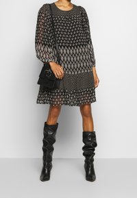 Svart mönstrad klänning med långa puffärmar, med en plisserad kjol och diamantformer. Bärs med knähöga svarta läderstövlar och en liten väska.