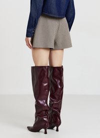 Stivali bordeaux al ginocchio in pelle lucida con design elegante e aderente e punta appuntita, abbinati a pantaloncini a vita alta a quadretti.