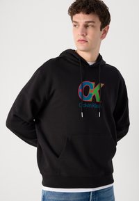 Fekete kapucnis pulóver elülső kenguruzsebbel, színes "OK" logóval és "Calvin Klein" felirattal. Puha anyagból készült.