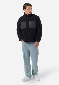 Črna fleece jakna z dvema črnima nalepkama za žep, visokim ovratnikom in sprednjim zadrgo. Občutek je svež, saj je oblečena z svetlo modrimi ohlapnimi kavbojkami in belimi supergah.