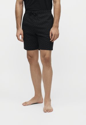 Homme portant un short noir à motifs avec cordon de serrage et une chemise noire, debout pieds nus devant un fond blanc uni.