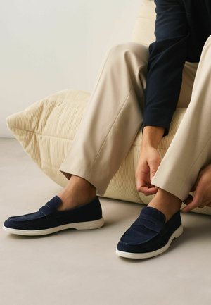 Osoba oblečená v béžových nohaviciach a námorníckomodrých slip-on topánkach si upravuje nohavice, zatiaľ čo sedí na svetlo béžovom textúrovanom vankúši.