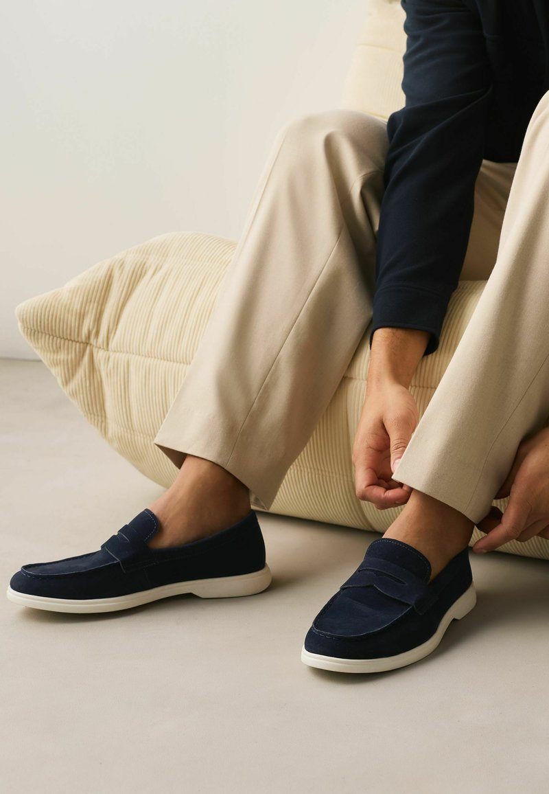 Isik, kes kannab beežikaid pükse ja tumesiniseid libisevaid kingi, kohandab istudes helebeeži tekstuuriga patja toetudes pükste säärt.