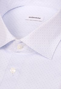 Gros plan d’une chemise habillée bleu clair à motifs, avec des boutons blancs et une étiquette de la marque seidensticker à l’intérieur du col.
