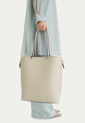 Ruka držící béžovou tašku na rameno s černými uchy, oblečená v světle modrém dlouhém kabátu a ladících širokých kalhotách s béžovými špičatými botami.