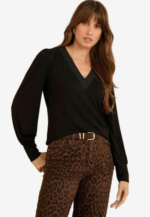 Femeie cu păr lung, castaniu, purtând un top negru cu decolteu în V și mâneci lungi, împreună cu pantaloni maro cu model leopard și o curea maro.