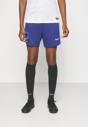 Person trägt blaue Nike-Fußballshorts mit Hahn-Emblem, lange schwarze Nike-Socken, schwarze Fußballschuhe und ein weißes Sporttrikot.