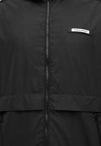Chaqueta acolchada negra con cremallera frontal, presenta una costura horizontal y un parche con el logo EA7 en la parte superior izquierda. Textura suave y diseño ligero.