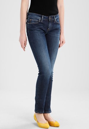 Personne portant un jean slim bleu foncé et des chaussures plates jaunes, debout devant un fond clair uni.