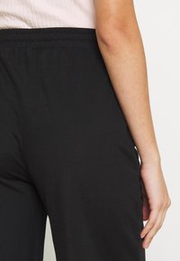 Pantalons noirs avec une taille élastique, en tissu texturé. Gros plan de l'arrière avec une main reposant sur la hanche.