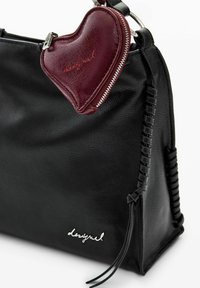 Geantă de mână din piele neagră cu detalii împletite, având un charm bordou în formă de inimă cu fermoar. Logo argintiu pe partea din față.