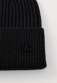 Beanie nero a maglia con texture a costine verticali, dotato di un piccolo logo "CK" nero. Materiale morbido con orlo pieghevole.