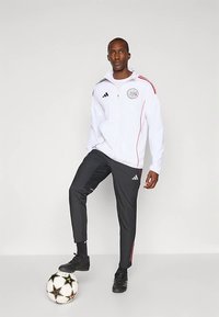 Fehér cipzáras dzseki fekete és piros díszítéssel, logó a mellkason, fekete nadrággal és fekete focicipővel párosítva, egy focilabda mellett állva.