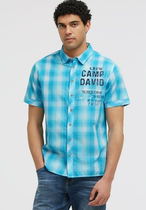 Junger Mann mit lockigem Haar, der ein blau kariertes Kurzarmhemd mit Textdruck und blaue Jeans vor einem einfarbigen Hintergrund trägt.