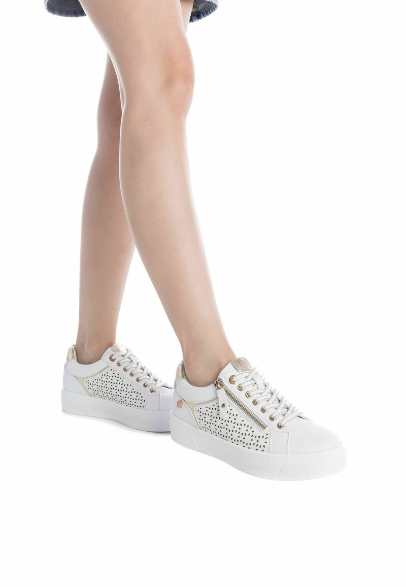 Zapatillas blancas con diseño perforado, detalles en dorado y cordones redondos. Presentan una suela plana y una cremallera en el lado exterior.