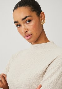 Beige textuur bovenstuk met hoge hals, met subtiele plooien. Gouden oorcuff met horizontale strepen, die de outfit aanvult.