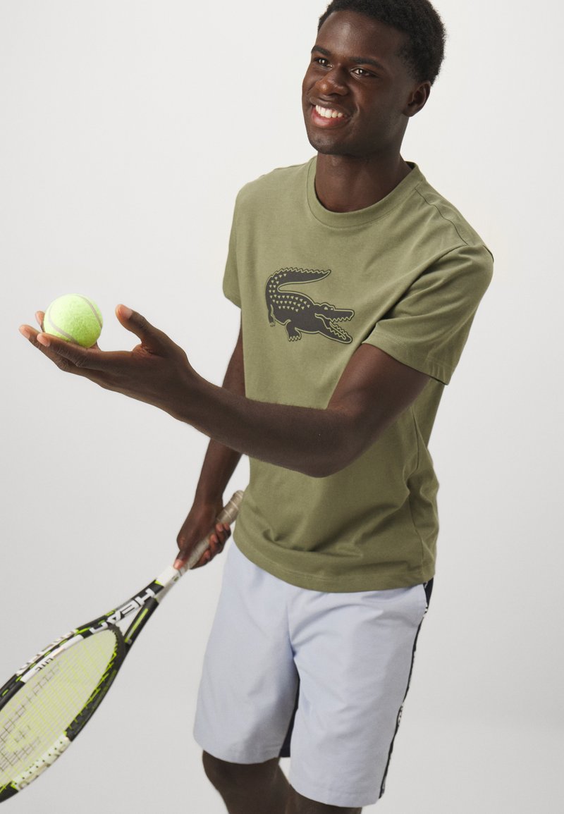 Olijfgroene t-shirt met korte mouwen en een zwart krokodillenlogo, lichtblauwe shorts, een tennisbal en een racket vasthoudend. Eenvoudig ontwerp.