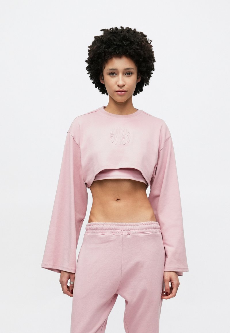 Šviesiai rožinė trumpa džemperio palaidinė su ilgomis rankovėmis ir siuvinėtais detalėmis. Derinama su derančiomis rožinėmis sportinėmis kelnėmis su elastiniu liemeniu.