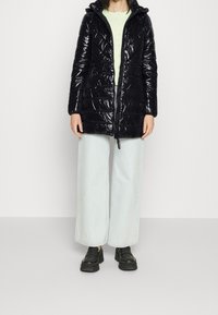 Czarna, błyszcząca kurtka puchowa z kapturem, z zapięciem na zamek, pionowymi szwami i bocznymi kieszeniami, noszona na jasnoszarym topie i szerokich dżinsach.