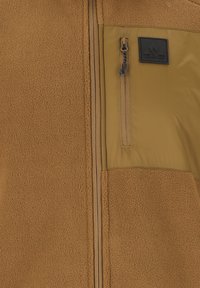 Ruskea fleece-takki, jossa on khakinvärinen nailon-yläosa. Takissa on vetoketju, vetoketjullinen rintatasku ja brändätty patch vasemmassa rinnassa.