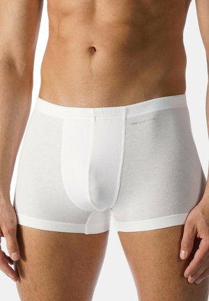 Männlicher Torso trägt schlichte weiße Baumwoll-Boxershorts, steht mit locker an den Seiten hängenden Armen vor einem hellen Hintergrund.