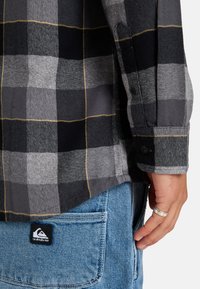 Gray and black plaid flannel shirt with yellow lines, featuring buttoned cuffs. Paired with blue denim jeans displaying logo tag.