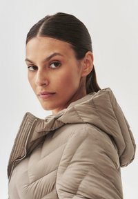 Vaalean beige puffer-takki hupulla, jossa on tikattu viisto kuviointi ja hopeavetoketju. Pehmeä pinta, suunniteltu lämpöä varten.