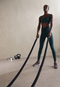 Egy zöld sportos öltözetbe öltözött személy fekete csata köteleket szorít az kezében, egy bézs padlón áll. Mögötte súlyzók vannak elrendezve. Az atlétikai cipők szürkék.