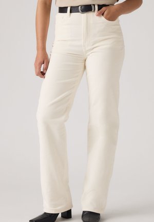 Persona con pantalones de pana blancos de talle alto, cinturón negro y zapatos negros de tacón, de pie frente a un fondo claro y liso.