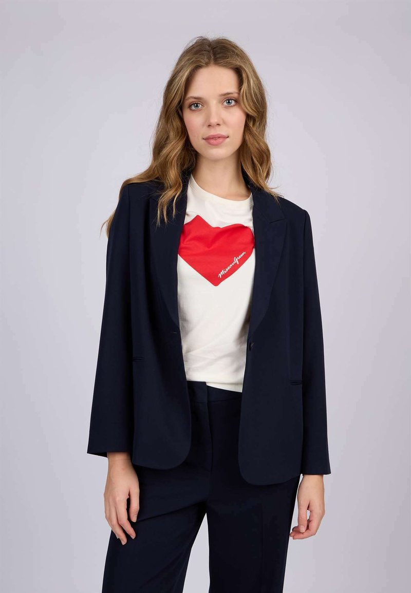 Blazer marine en tissu lisse, un bouton avec revers crantés, associé à un t-shirt blanc avec un graphisme de cœur rouge.