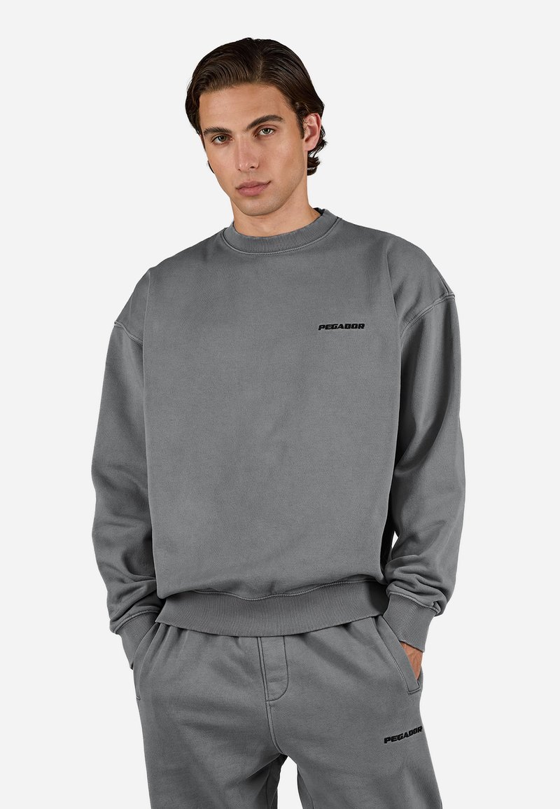 Grauer Sweatshirt aus weichem Material, mit Rundhalsausschnitt und gerippten Bündchen. Zeigt schwarz das "PEGADOR"-Logo auf der Brust.