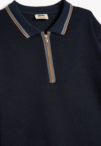 Camisa polo azul marino con una tapeta con cremallera, textura acanalada y rayas contrastantes en color beige y blanco a lo largo del cuello y la tapeta.