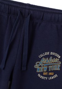 Marineblauwe sweatpants met een elastische tailleband en trekkoord, met een witte en gouden bedrukte logo dat "Athletic New York" zegt.