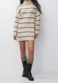 Femme portant une robe-pull à col roulé beige avec des rayures horizontales bleu marine, des bottes marron montant jusqu'aux genoux et des chaussettes grises détendues sur un fond blanc.