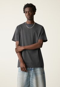 Dunkelgraues Baumwoll-T-Shirt mit kurzen Ärmeln, rundem Ausschnitt und lockerem Schnitt. Ermöglicht eine silberne Kettenkette und hellblaue Denim-Shorts.
