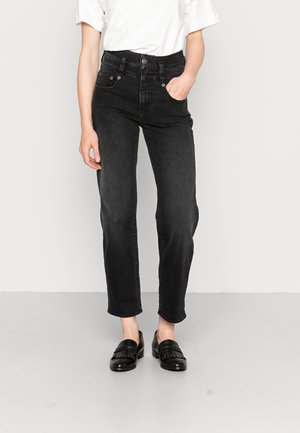 Frau trägt hoch taillierte schwarze Jeans mit geraden Beinen, eine Hand in der Tasche, weißes Oberteil und schwarze Loafers, steht auf grauem Boden.